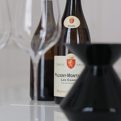 A close-up shot of two bottles of Burgundy white wine: a 2020 Puligny-Montrachet from Domaine Nudant, with a blurred wine glass in the foreground and an opaque green spittoon. The image highlights the professional tasting methodology taught at Sensation V